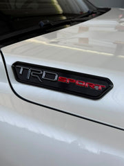 Close-up of a white car door badge with a black TRD SPORT emblem, photographed in a garage setting.
