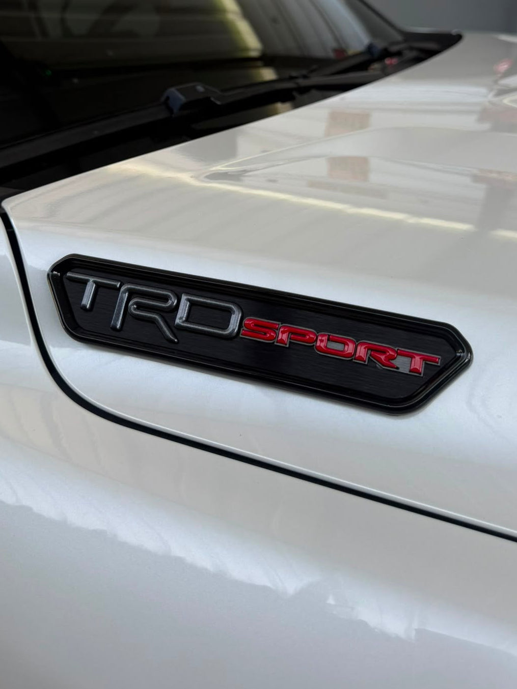 Close-up of a white car door badge with a black TRD SPORT emblem, photographed in a garage setting.